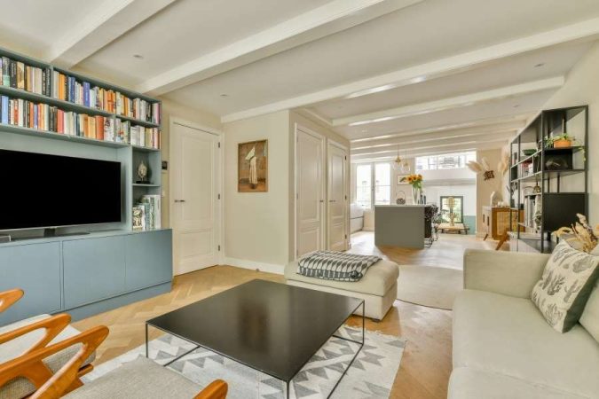 Un salon ouvert avec poutres apparentes blanches, bibliothèque bleue intégrée et mobilier épuré illustrant une décoration minimaliste élégante et lumineuse.