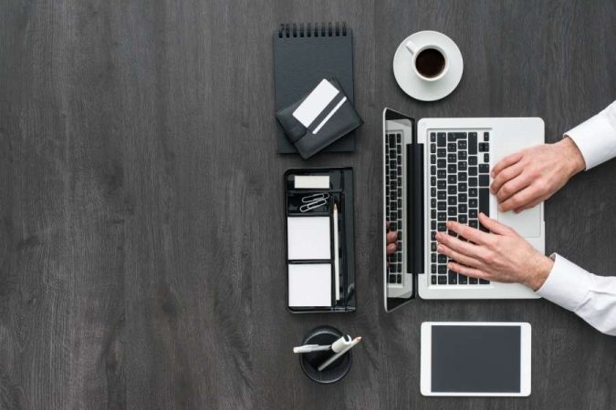 Vue de dessus d'un bureau en bois sombre où un professionnel en chemise blanche tape sur un ordinateur portable à côté d'une tasse de café et d'une tablette.