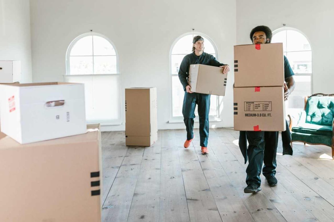 Deux déménageurs en uniforme portant des cartons dans un grand salon lumineux. Découvrez combien coûte un déménagement fait par des professionnels.