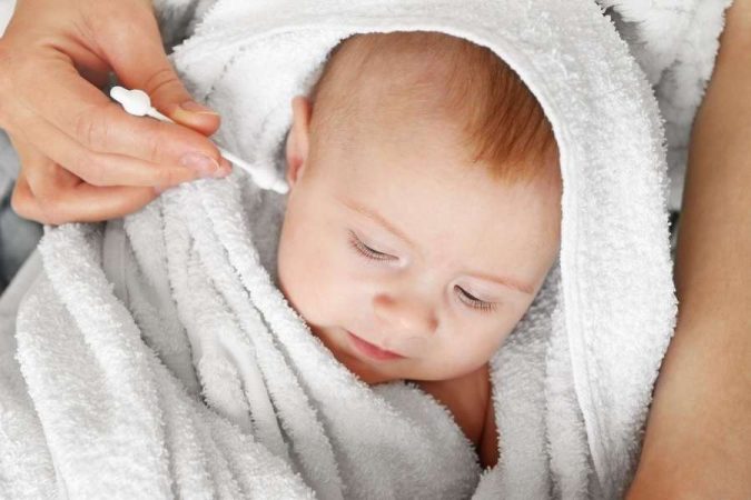 Un bébé emmitouflé dans une serviette blanche, les yeux fermés, pendant qu'un parent s'apprête à nettoyer les oreilles d'un bébé.