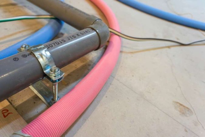 Gros plan sur une canalisation grise fixée par un collier de serrage métallique, avec un tube rose gainé visible en arrière-plan.